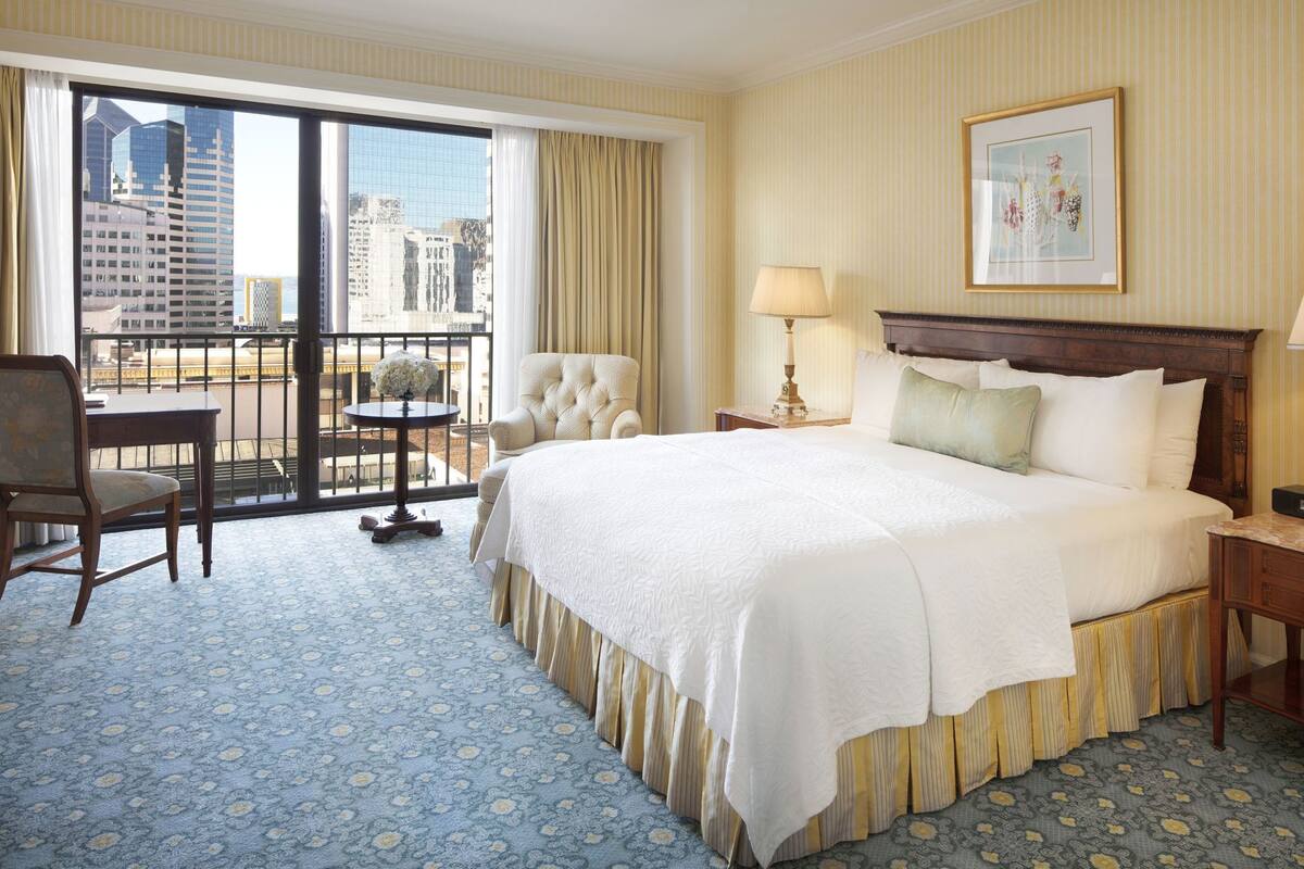 Quarto de hotel com cama grande, uma cadeira, uma mesa, abajur e um quadro na parede.