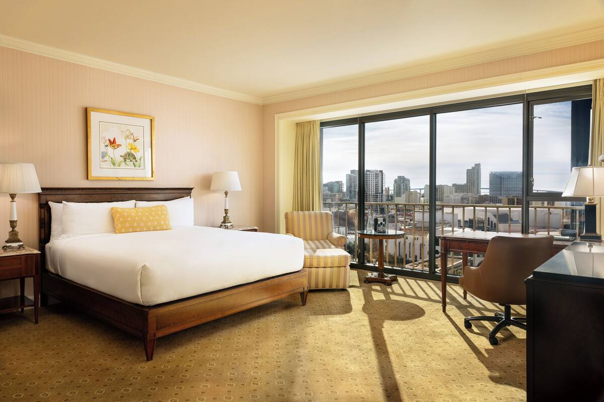 Quarto de hotel com uma cama grande, uma escrivaninha, uma cadeira e vista para a cidade através de amplas janelas.