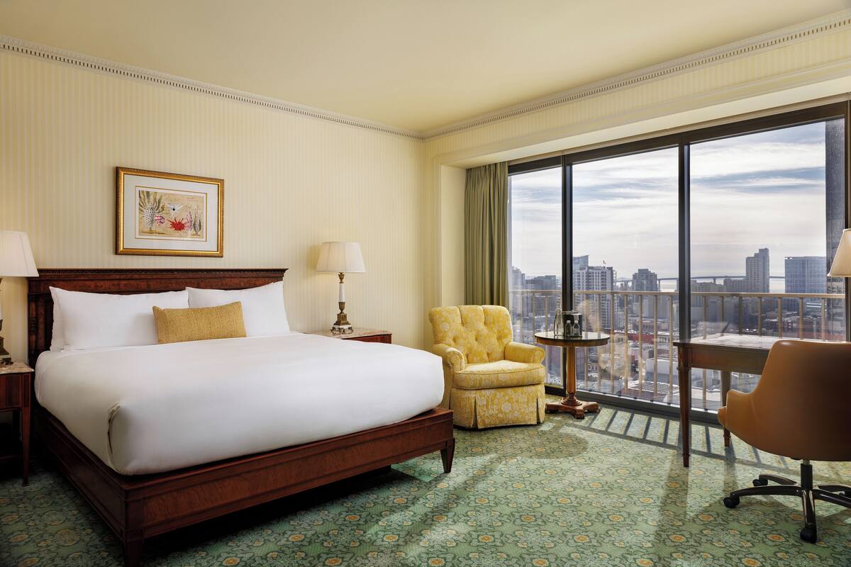 Quarto de hotel com uma cama grande, uma cadeira, uma mesinha e vista da cidade através de grandes janelas.