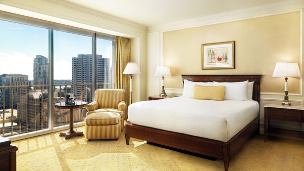 A hotel room with a large bed, two bedside tables, a chair, and a view of the city through large windows. - Westgate Hotel (San Diego)