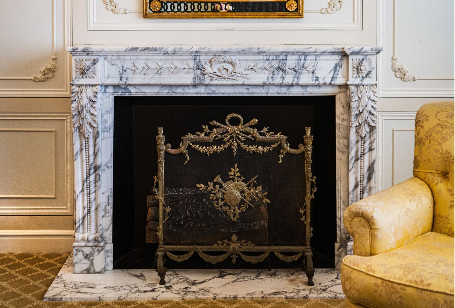 A fireplace with an ornate metal grate, a marble mantle, and a gold chair to the right.