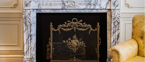 A fireplace with an ornate metal grate, a marble mantle, and a gold chair to the right.
