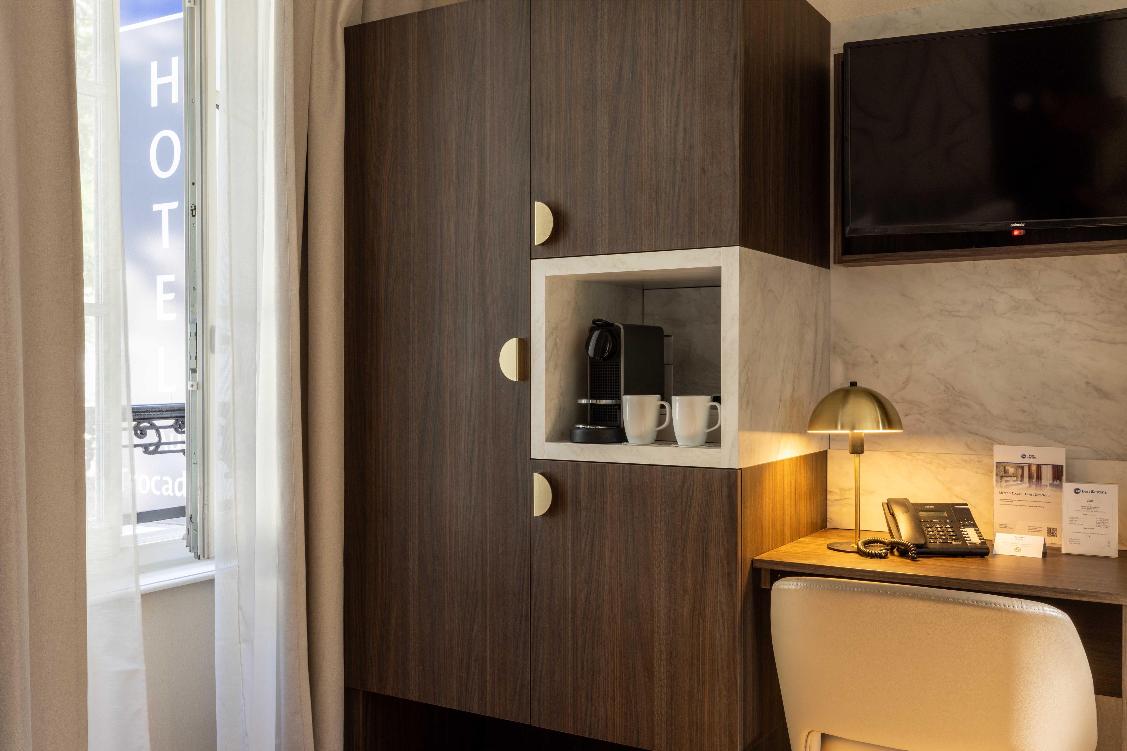 Ein Hotelzimmer mit Schreibtisch, einer Kaffeemaschine, einem Telefon und einem an der Wand befestigten Fernseher.