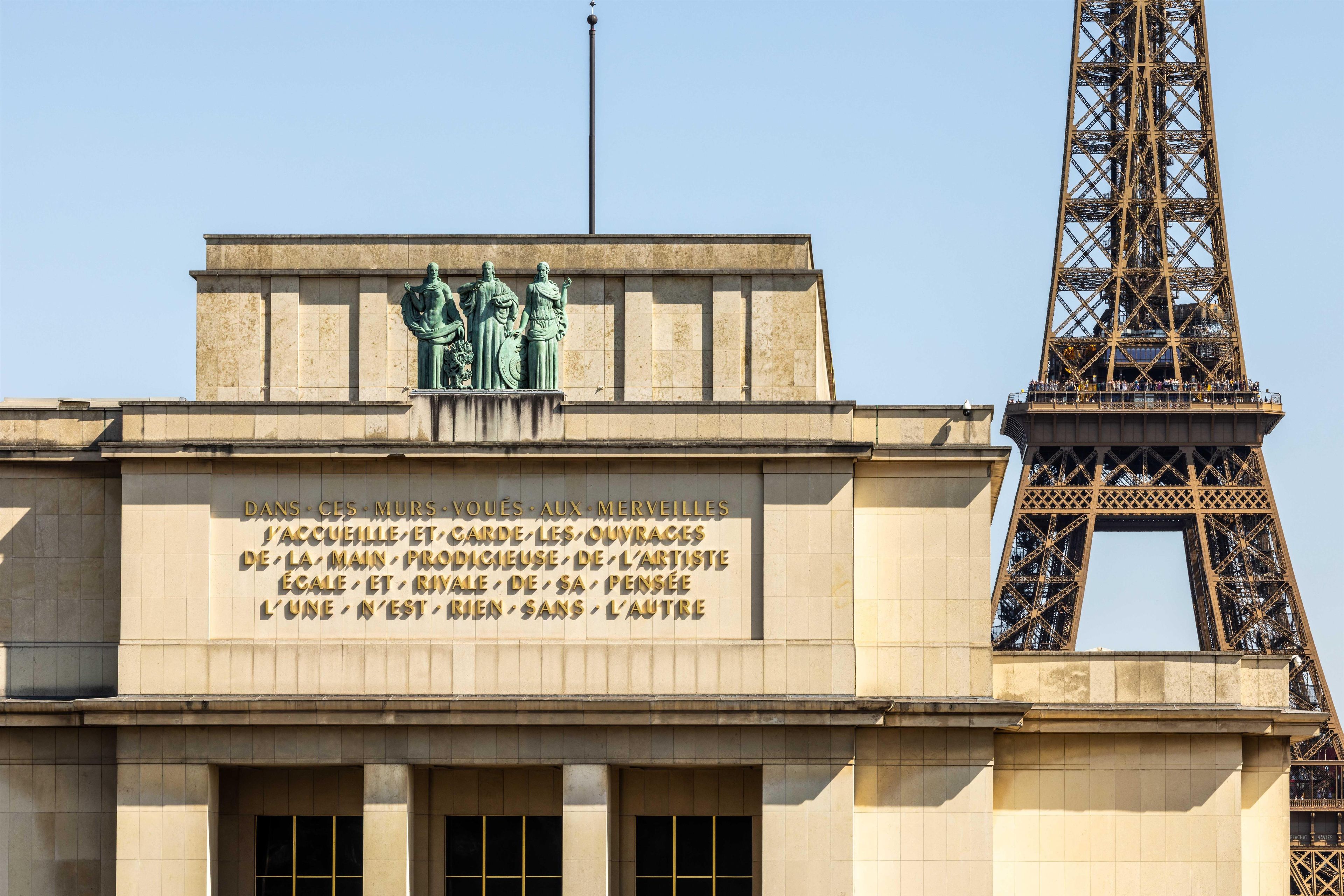 Fachada de un edificio con un gran letrero en francés, una estatua de tres figuras y la Torre Eiffel al fondo.