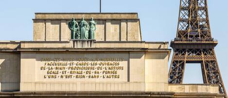 Fachada de un edificio con un gran letrero en francés, una estatua de tres figuras y la Torre Eiffel al fondo.