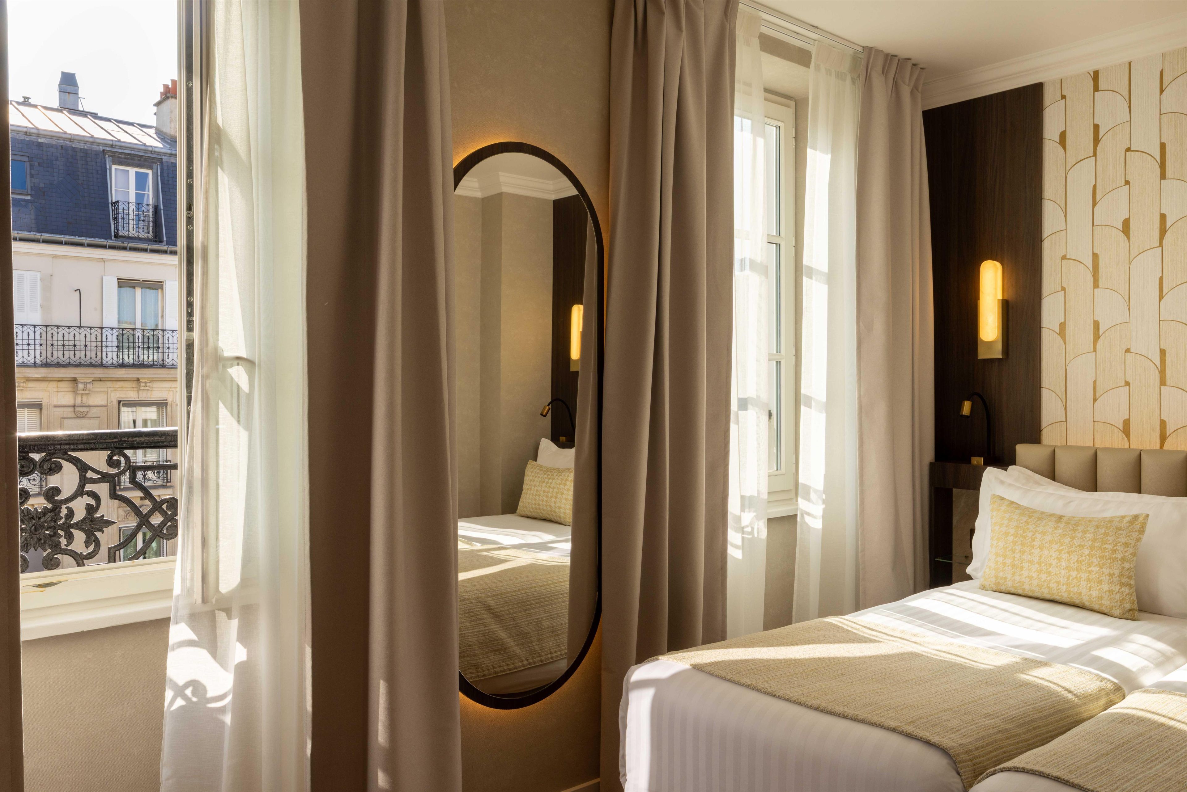A hotel room with a bed, a bedside table, a mirror, and a view of the outside.