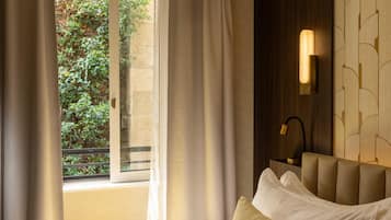 Quarto de hotel com cama, janela com cortinas, luminária na parede e vista para o verde lá fora.