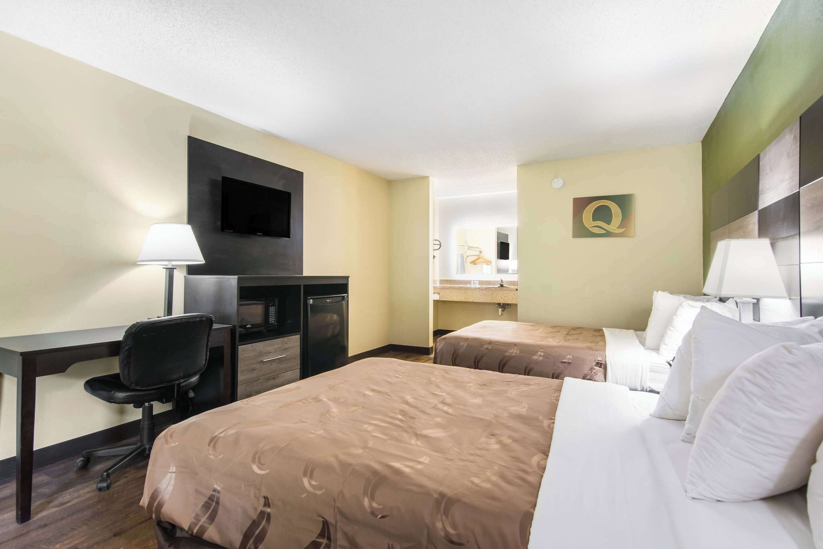 A hotel room with two beds, a desk, a chair, a TV, and a bathroom visible in the background.