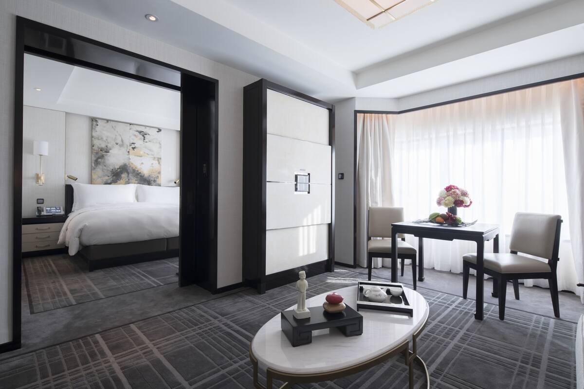 Una habitación de hotel moderna con una cama grande, un escritorio con un florero y vistas a la ciudad a través de amplias ventanas.