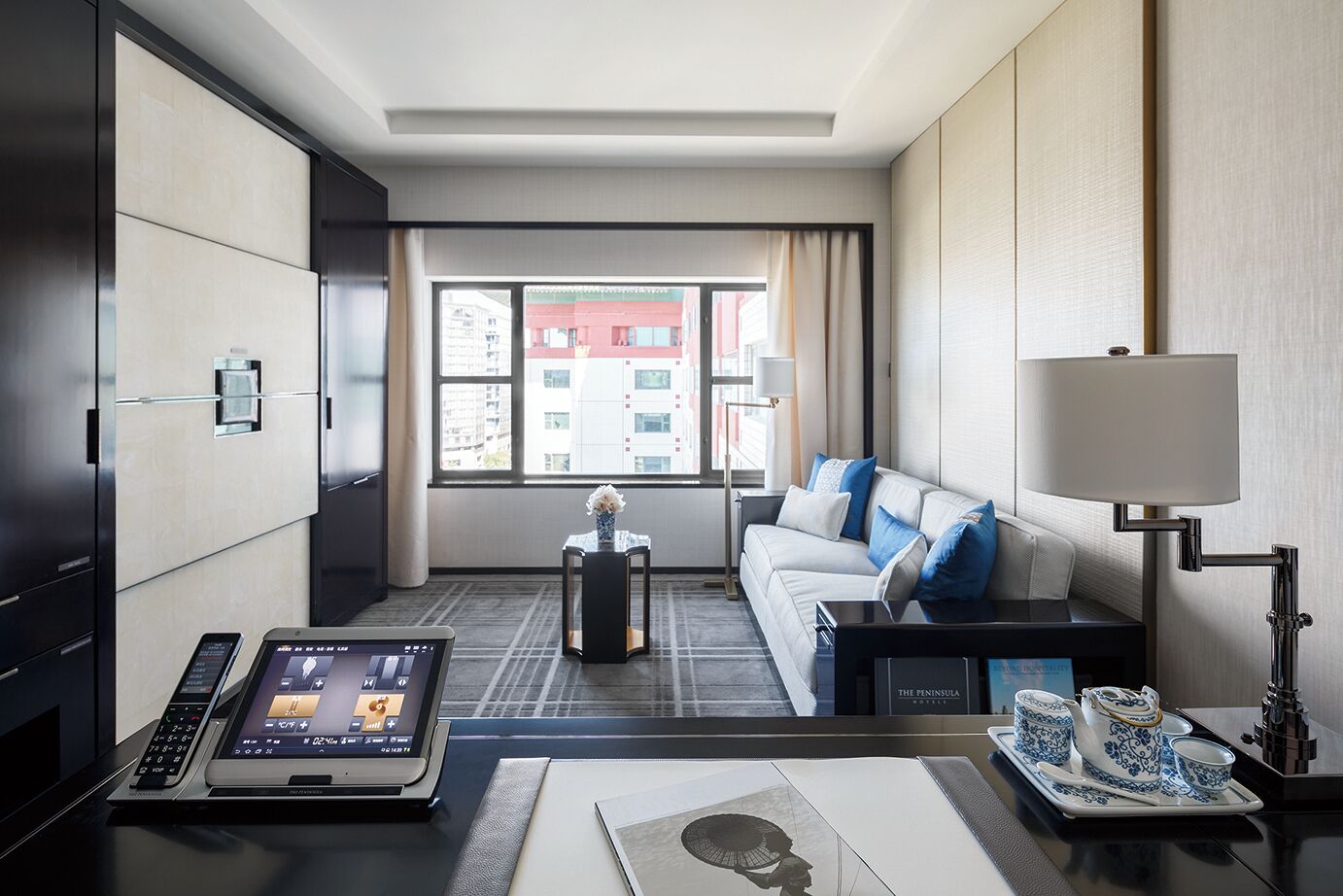 A modern hotel room with a sofa, a desk with a laptop, a bedside table with a lamp, and a view of the city through large windows.