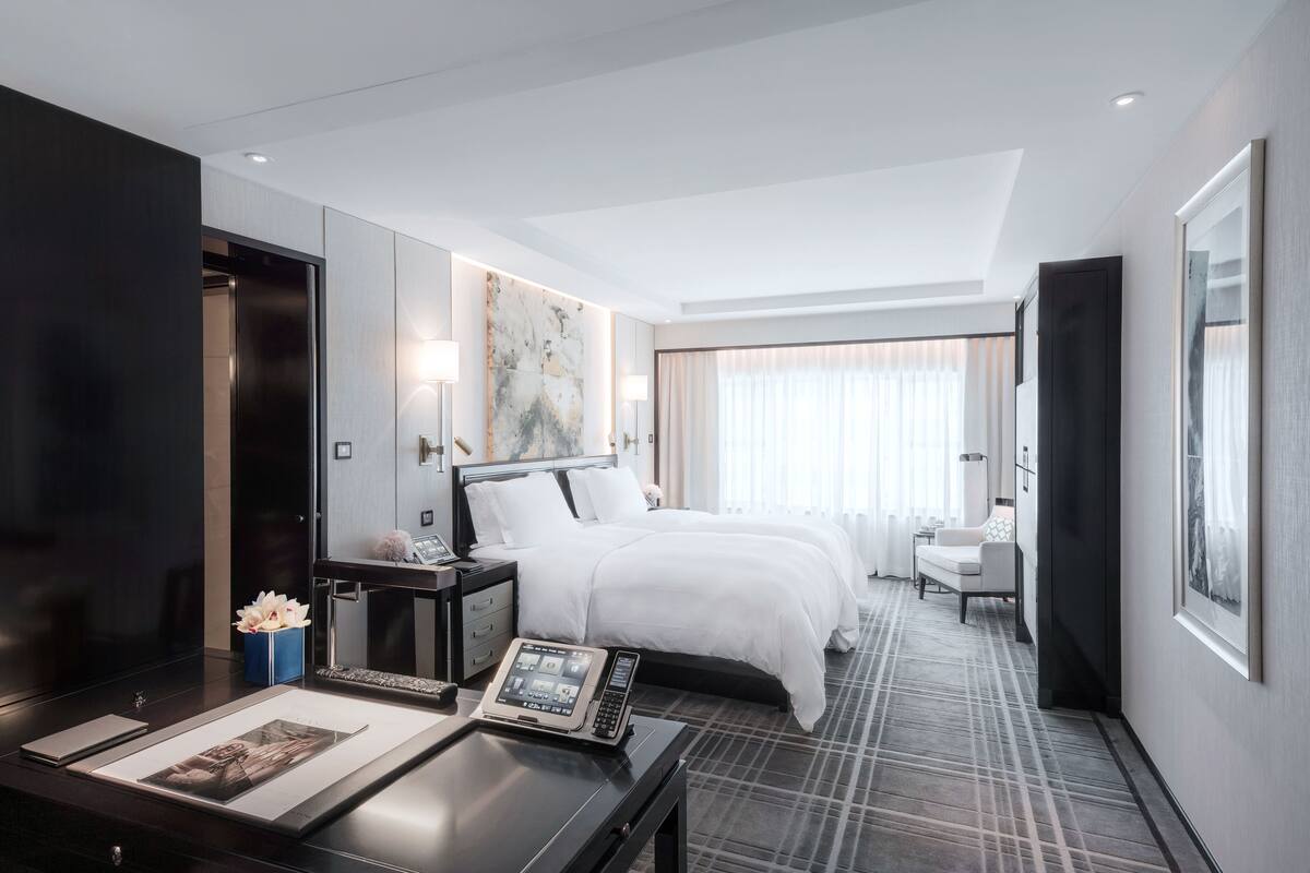 Una habitación de hotel moderna con una cama grande, un escritorio con una computadora portátil, una mesita de noche con una lámpara y un ventanal con cortinas.