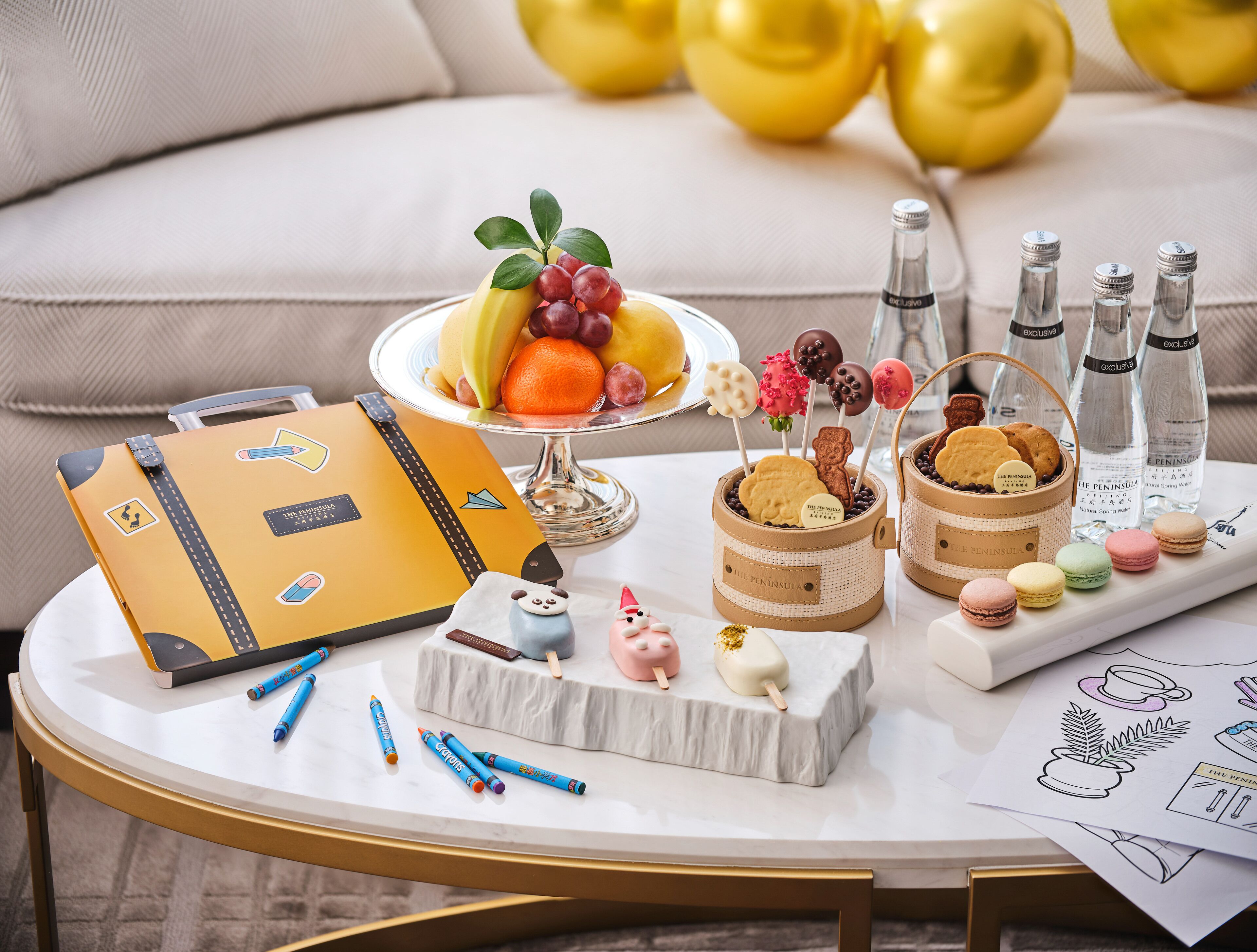 Une table avec une valise, un assortiment de fruits, des biscuits, des macarons et des bouteilles d’eau.