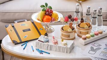 Une table avec une valise, un assortiment de fruits, des biscuits, des macarons et des bouteilles d’eau.