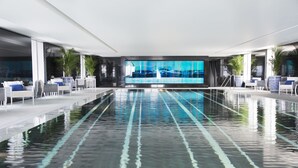 Piscina interior moderna con marcas de carriles bien definidas, rodeada de sillones de descanso y plantas en macetas.