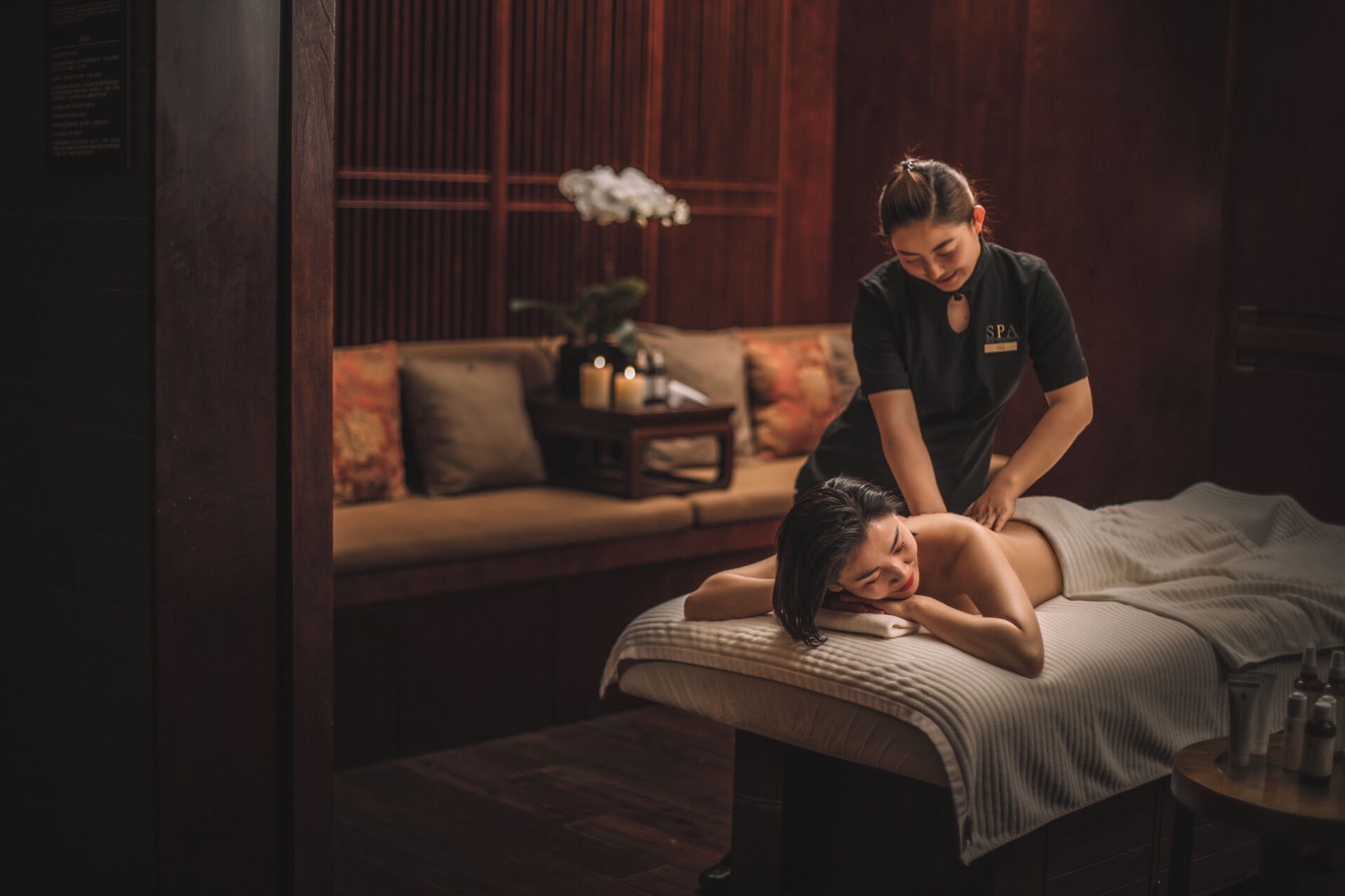 Eine Massagetherapie in einem schwach beleuchteten Raum mit Holzvertäfelung, einer Couch und einem kleinen Tisch mit Kerzen.
