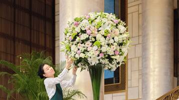 A person arranging a large floral centerpiece in a lobby with columns and a staircase.