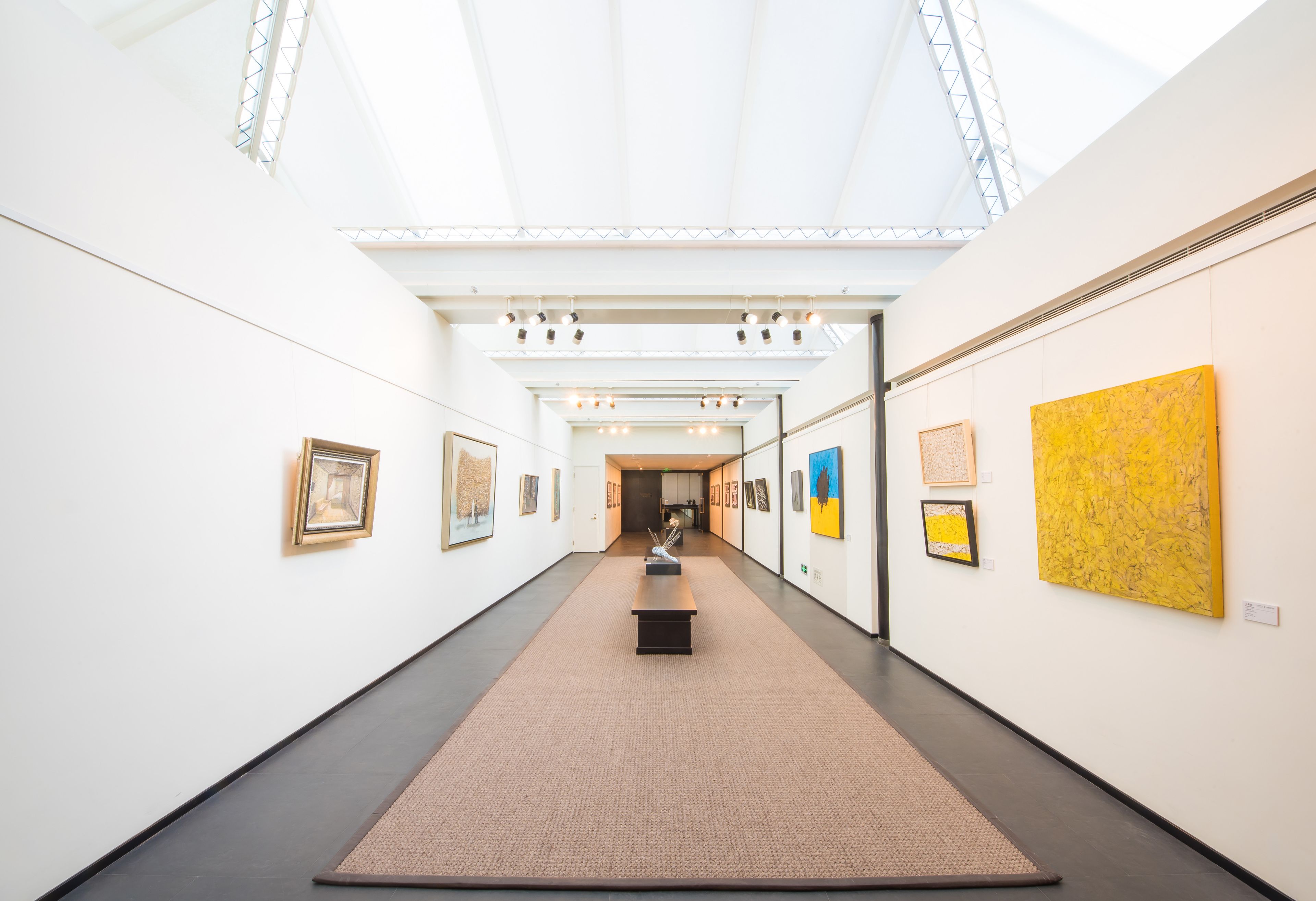 A modern art gallery with a central walkway, framed artworks on the walls, and a skylight ceiling.