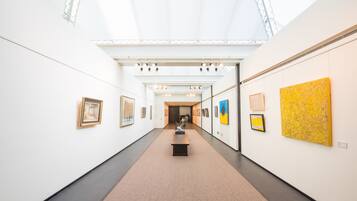 A modern art gallery with a central walkway, framed artworks on the walls, and a skylight ceiling.