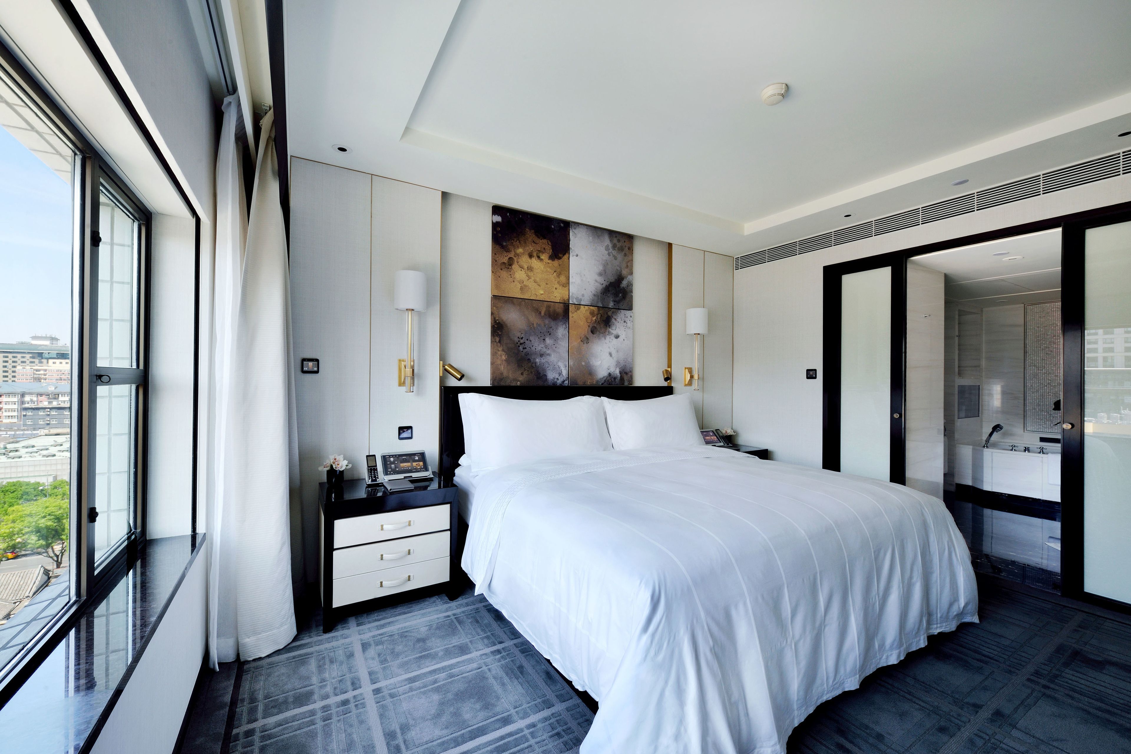 Una habitación de hotel moderna con una cama grande, una mesita de noche con teléfono y lámpara, vistas a la ciudad a través de un ventanal amplio y un baño con espejo y lavamanos.