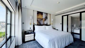 Una habitación de hotel moderna con una cama grande, una mesita de noche con teléfono y lámpara, vistas a la ciudad a través de un ventanal amplio y un baño con espejo y lavamanos.