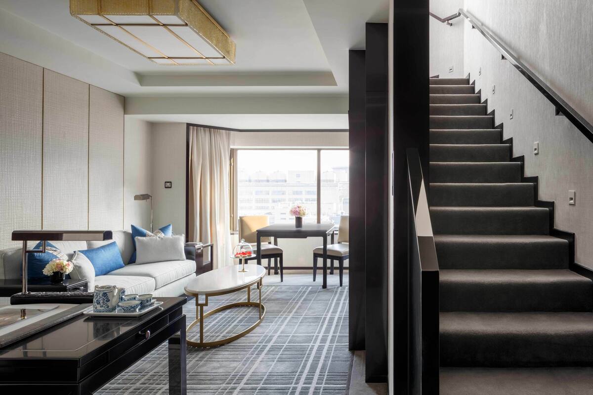 Una habitación de hotel moderna con sofá, mesa de centro y zona de comedor. Una escalera conduce a un piso superior.