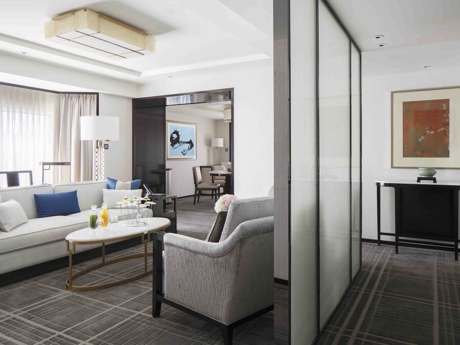 A modern hotel room with a sofa, armchair, coffee table, and a dining area.