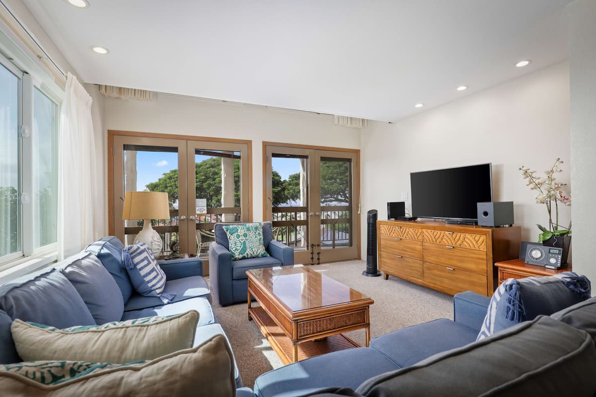 A spacious living room with a sofa, a coffee table, and a television. There are large windows and a balcony visible.
