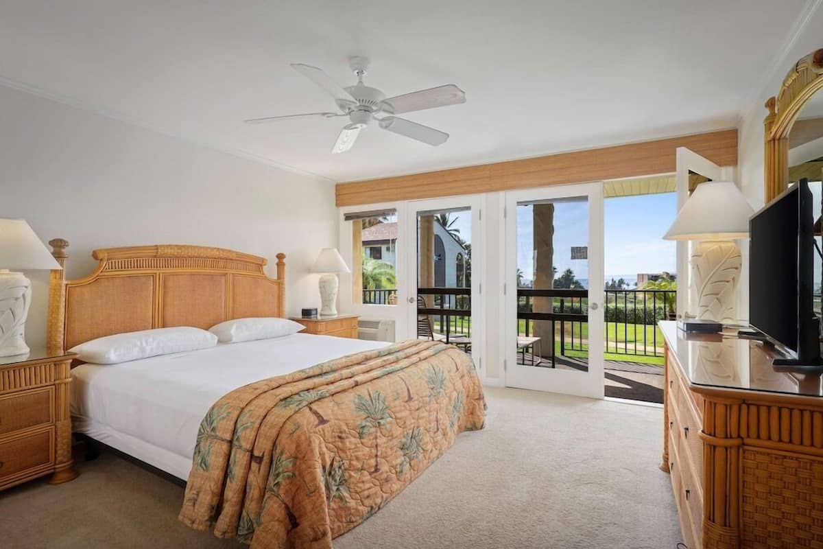 A bedroom with a large bed, a ceiling fan, a TV, and a balcony with a view.