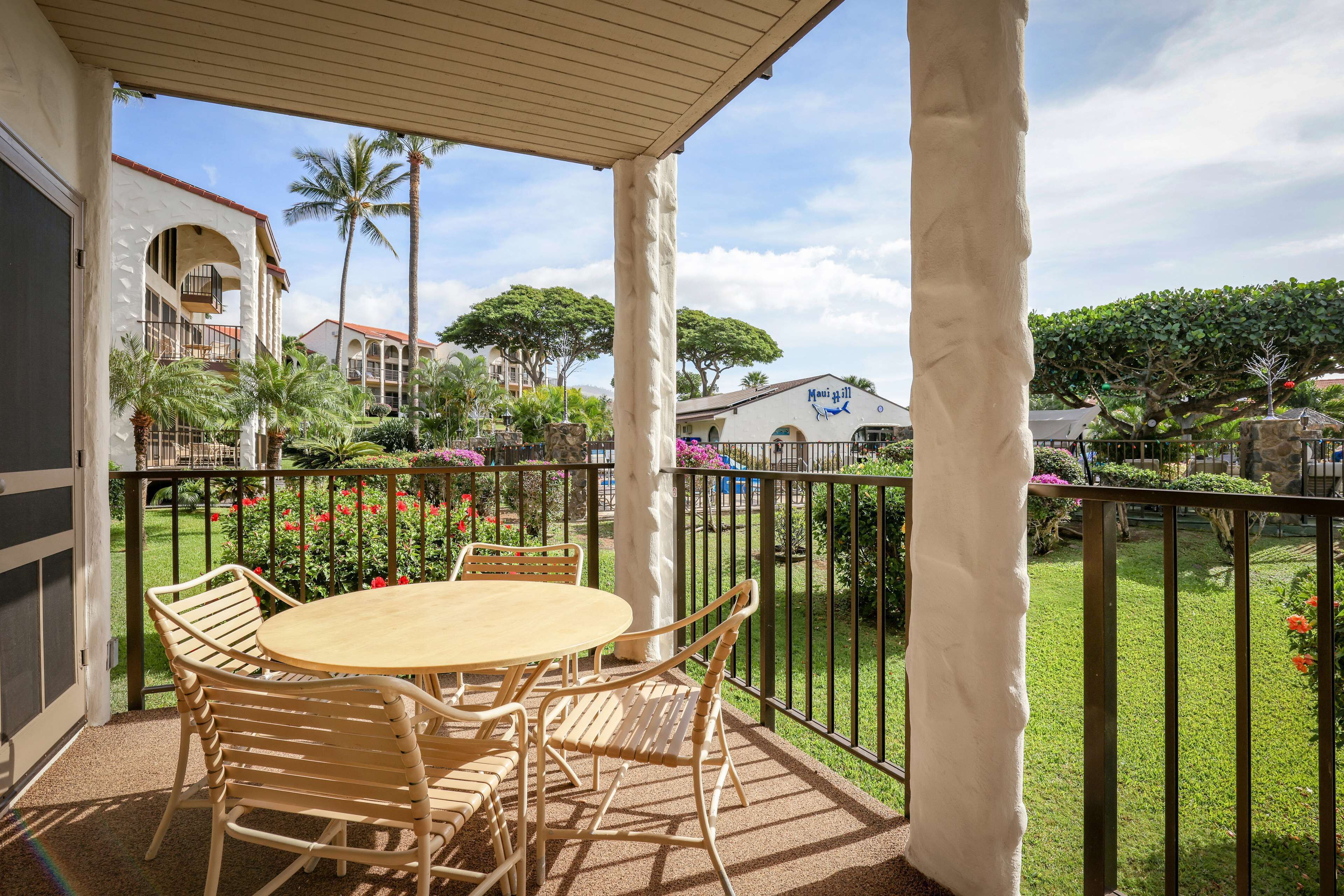 A covered outdoor area with a round table and chairs, surrounded by a metal fence, overlooking a garden with palm trees and a building labeled "Maui Grill."