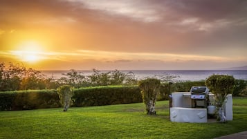 A grassy outdoor area with a barbecue grill and a view of the ocean at sunset.
