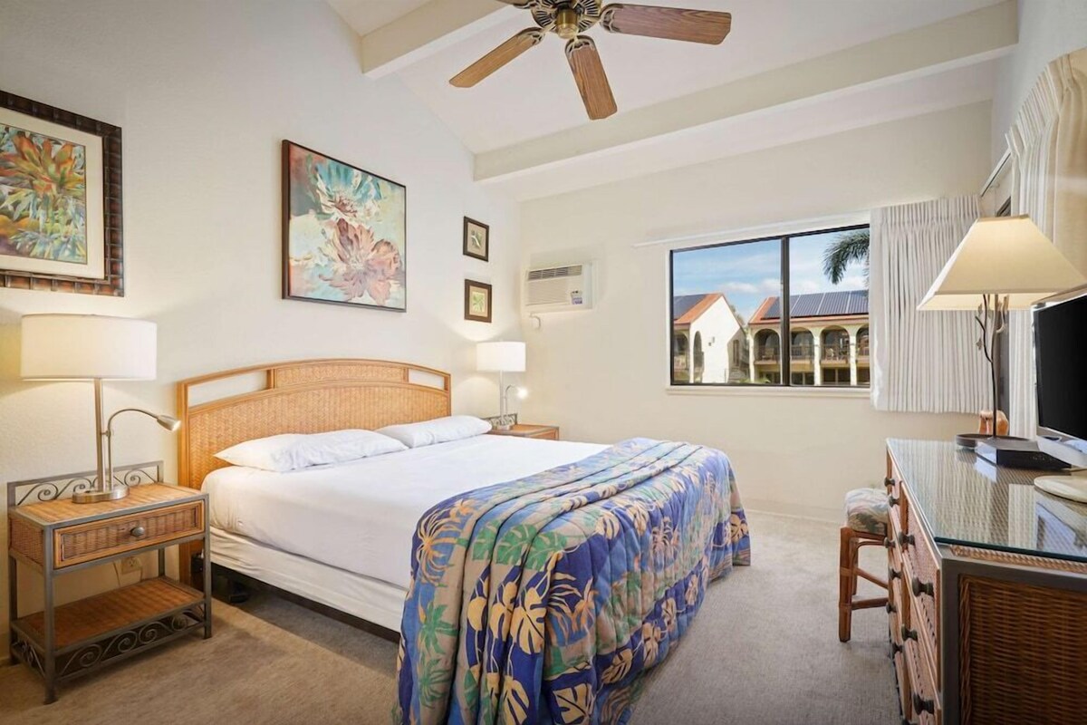 A hotel room with a bed, bedside tables, a desk, a chair, a ceiling fan, and a window with a view of buildings.