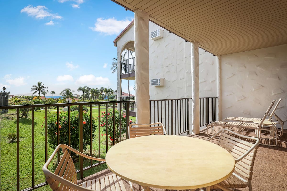 A covered outdoor seating area with a table and chairs, overlooking a well-maintained garden and a building with a balcony.