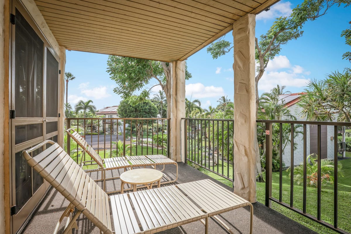A covered outdoor area with two lounge chairs and a small table, surrounded by a metal railing and a view of a garden and buildings.