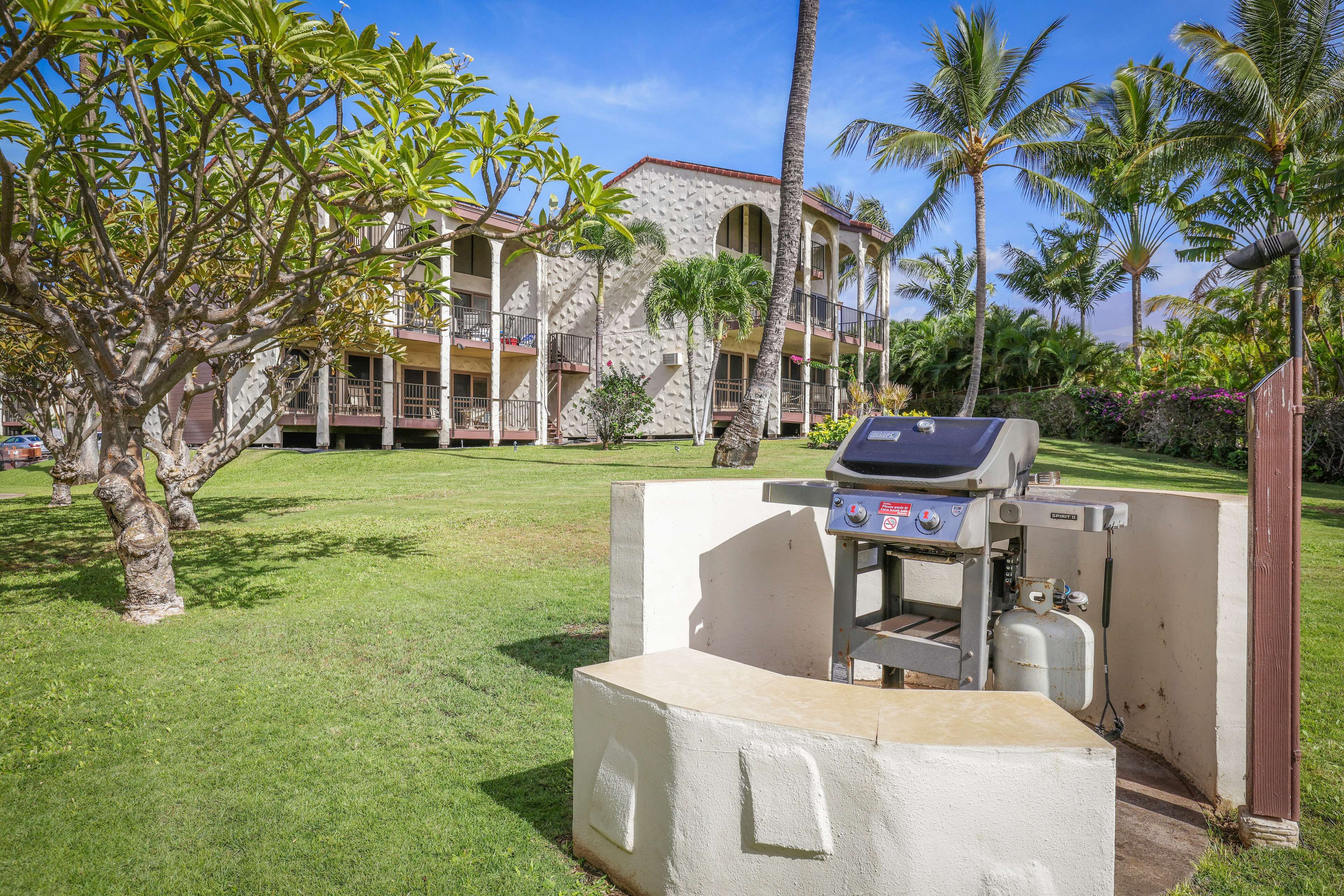 An outdoor grill area with a concrete structure, surrounded by palm trees and a building with balconies in the background.