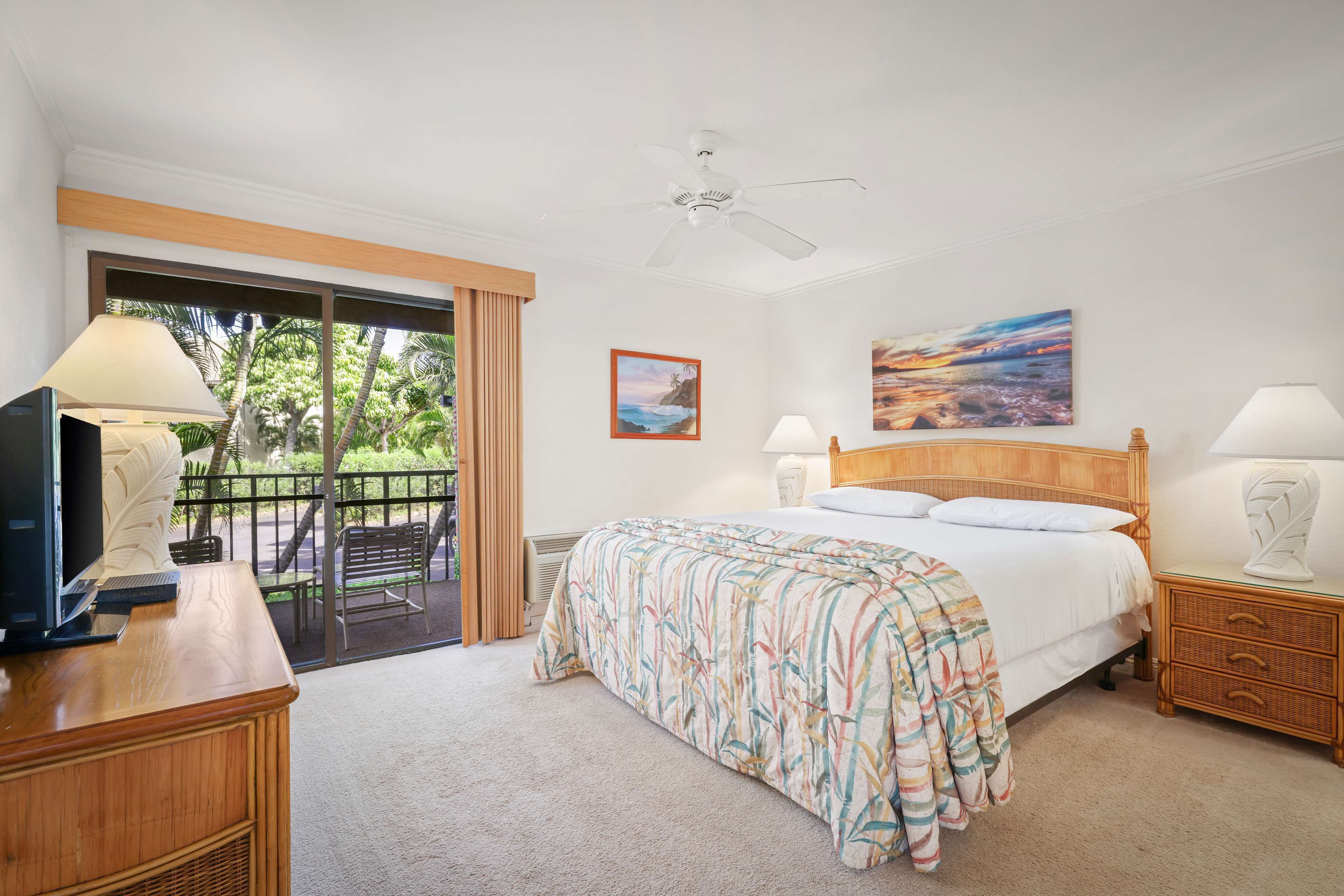 Une chambre d’hôtel comprenant un lit, un bureau avec un ordinateur, une télévision, un balcon avec vue sur les palmiers et un ventilateur de plafond.