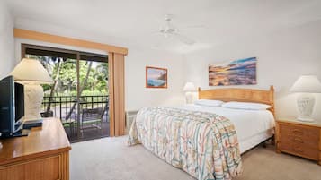Et hotelværelse med en seng, et skrivebord med en computer, et fjernsyn, en balkon med udsigt til palmer og en loftventilator.