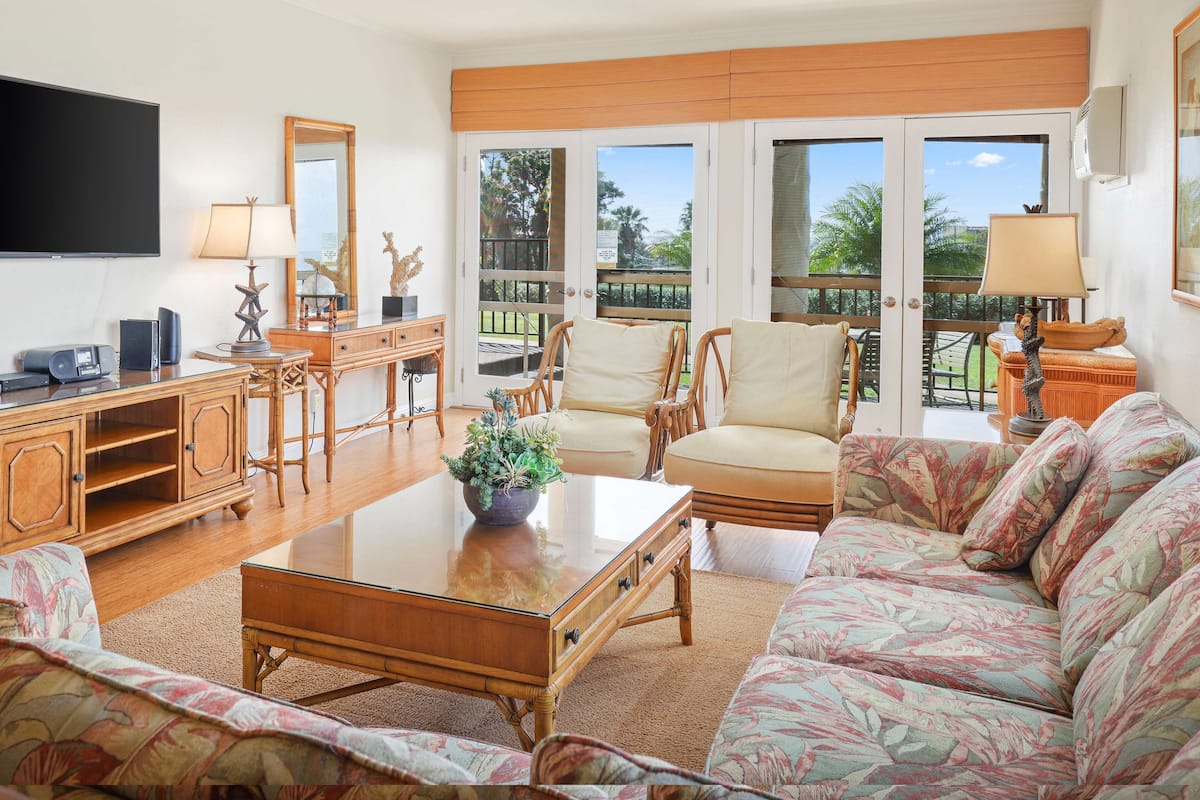 A living room with a sofa, coffee table, and chairs, a television, and a balcony with a view of palm trees.