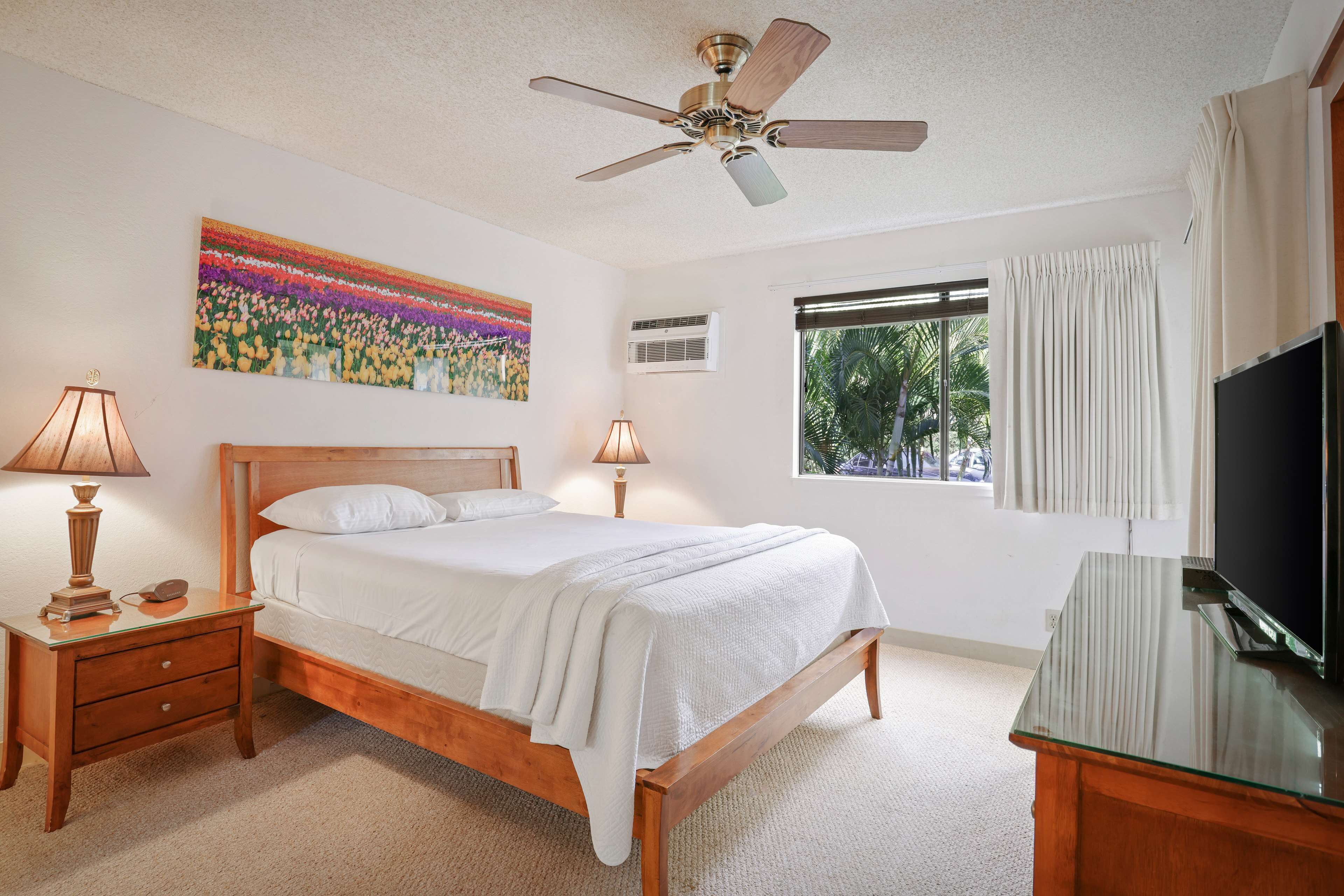 A bedroom with a wooden bed, two bedside tables with lamps, a ceiling fan, a window with curtains, and a TV on a glass table.