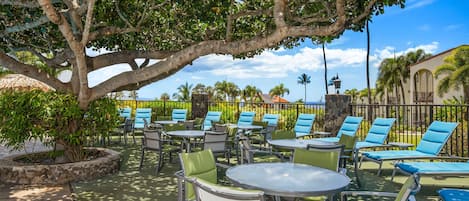 An outdoor seating area with round tables and chairs, surrounded by a stone wall and palm trees.