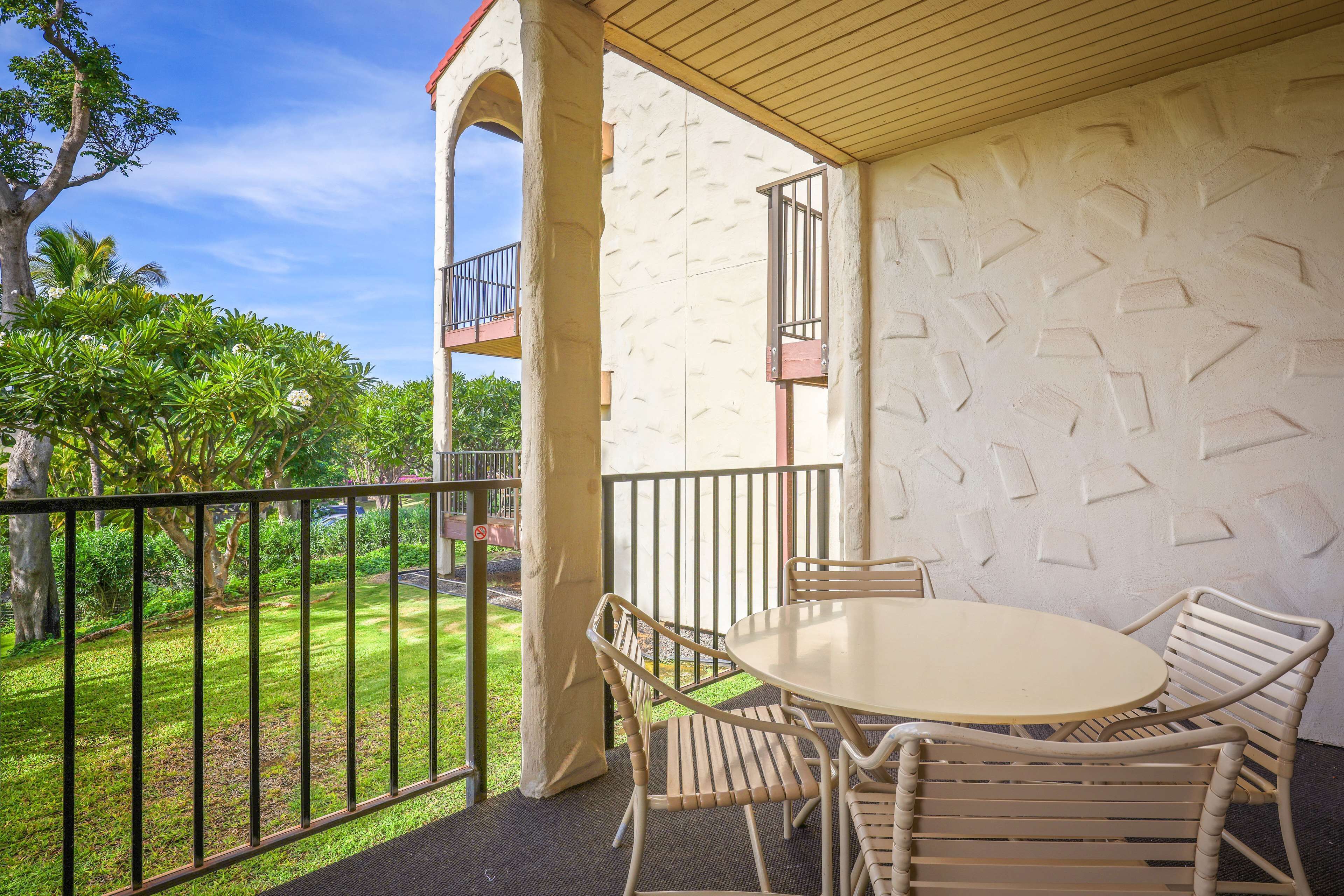 A covered outdoor seating area with a round table and chairs, a balcony with a railing, and a view of a green lawn and trees.