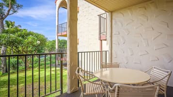A covered outdoor seating area with a round table and chairs, a balcony with a railing, and a view of a green lawn and trees.