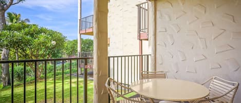 A covered outdoor seating area with a round table and chairs, a balcony with a railing, and a view of a green lawn and trees.