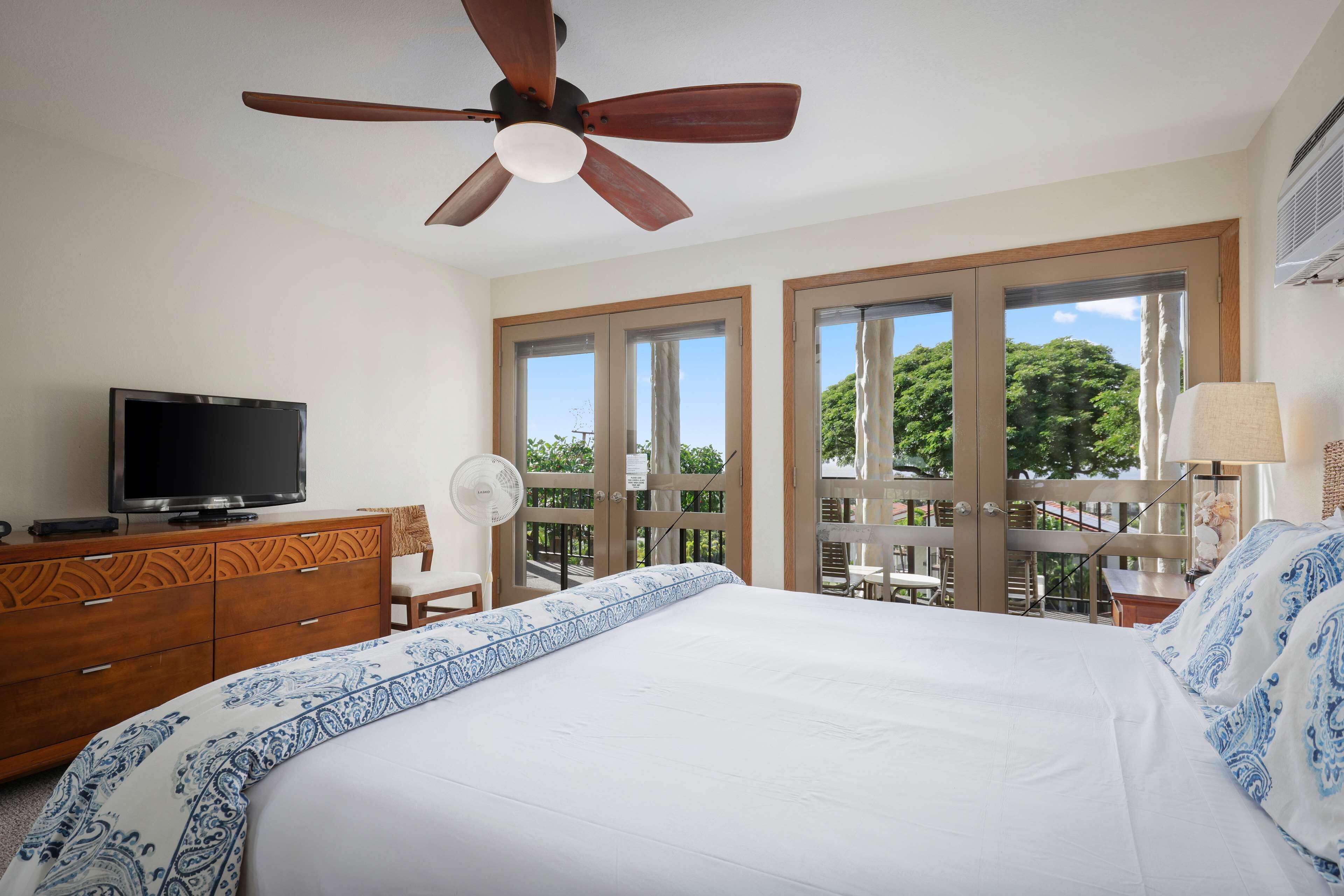 A bedroom with a bed, a TV on a dresser, a ceiling fan, and large windows.