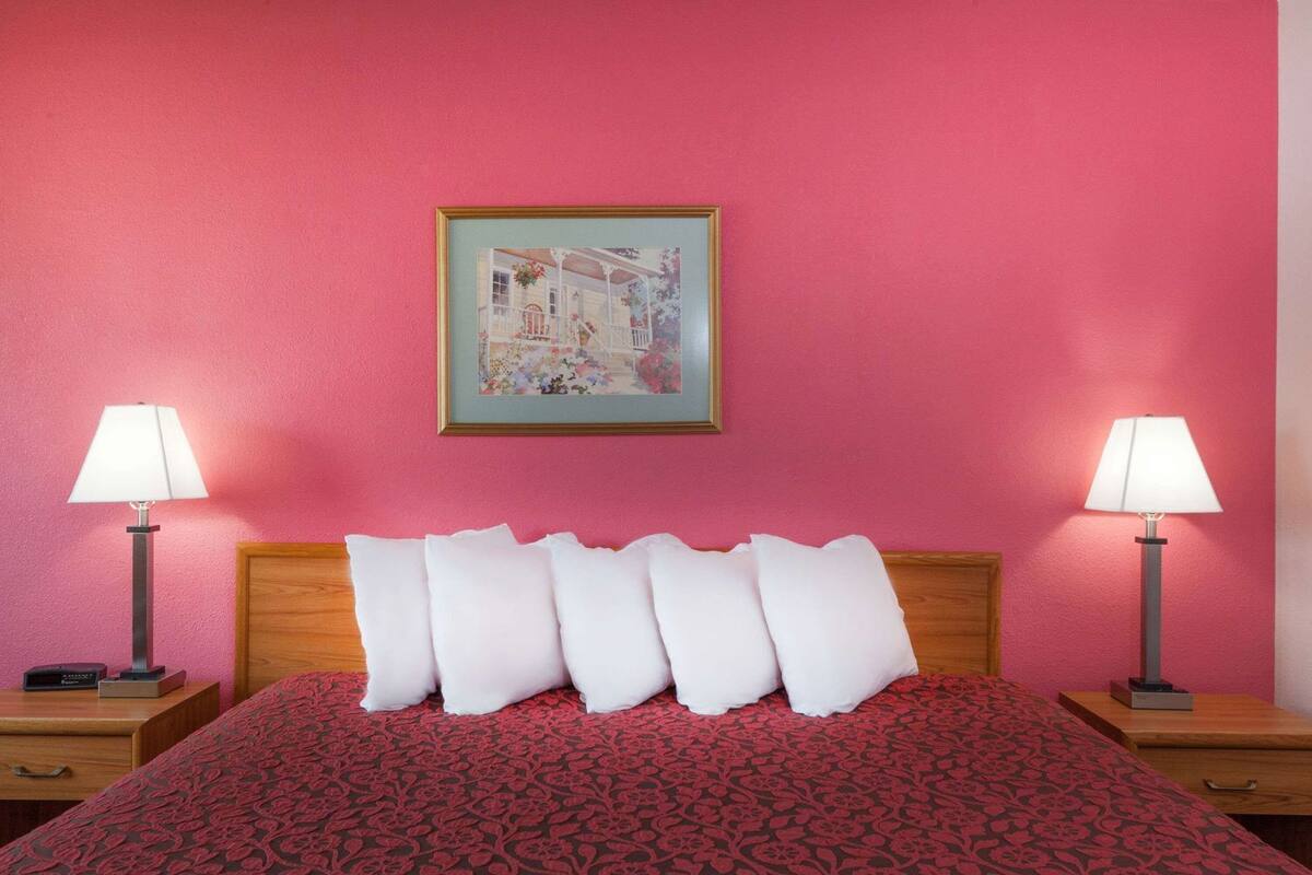 Une chambre d’hôtel avec un mur rose, un tableau encadré, un lit avec des oreillers blancs et deux lampes de chevet.