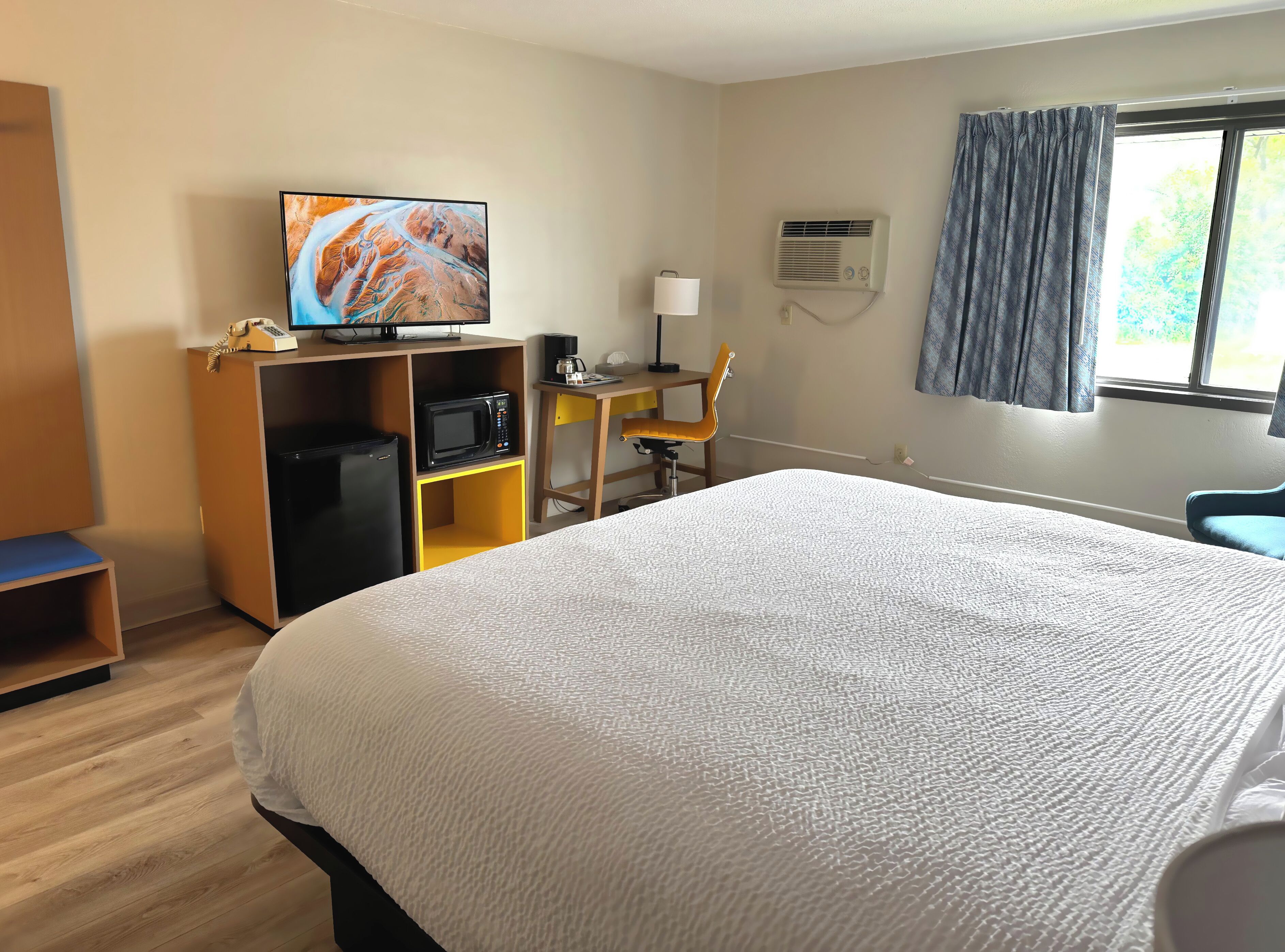 Quarto de hotel com uma cama grande, televisão em uma prateleira de madeira, escrivaninha com cadeira, micro-ondas e ar condicionado.