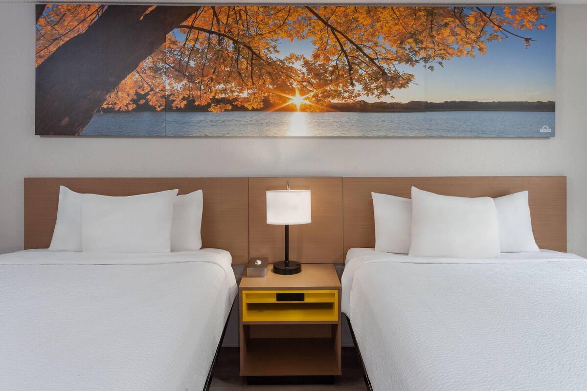 Une chambre d’hôtel avec deux lits, une table de chevet avec une lampe et un grand tableau représentant un coucher de soleil sur un lac.
