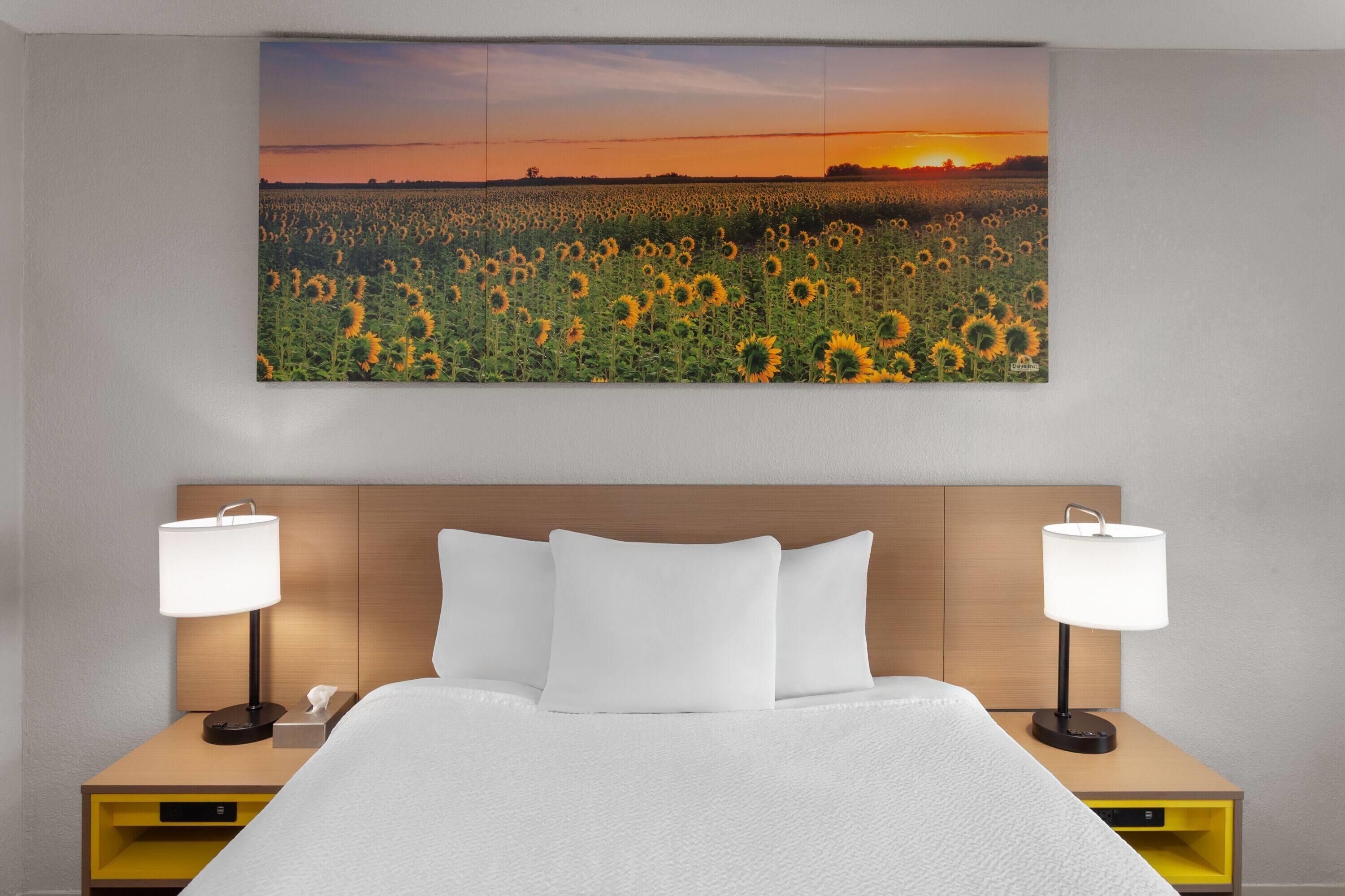 A hotel room with a large bed, two bedside tables with lamps, a wooden headboard, and a sunset sunflower field artwork on the wall.