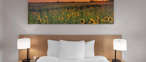 A hotel room with a large bed, two bedside tables with lamps, a wooden headboard, and a sunset sunflower field artwork on the wall.