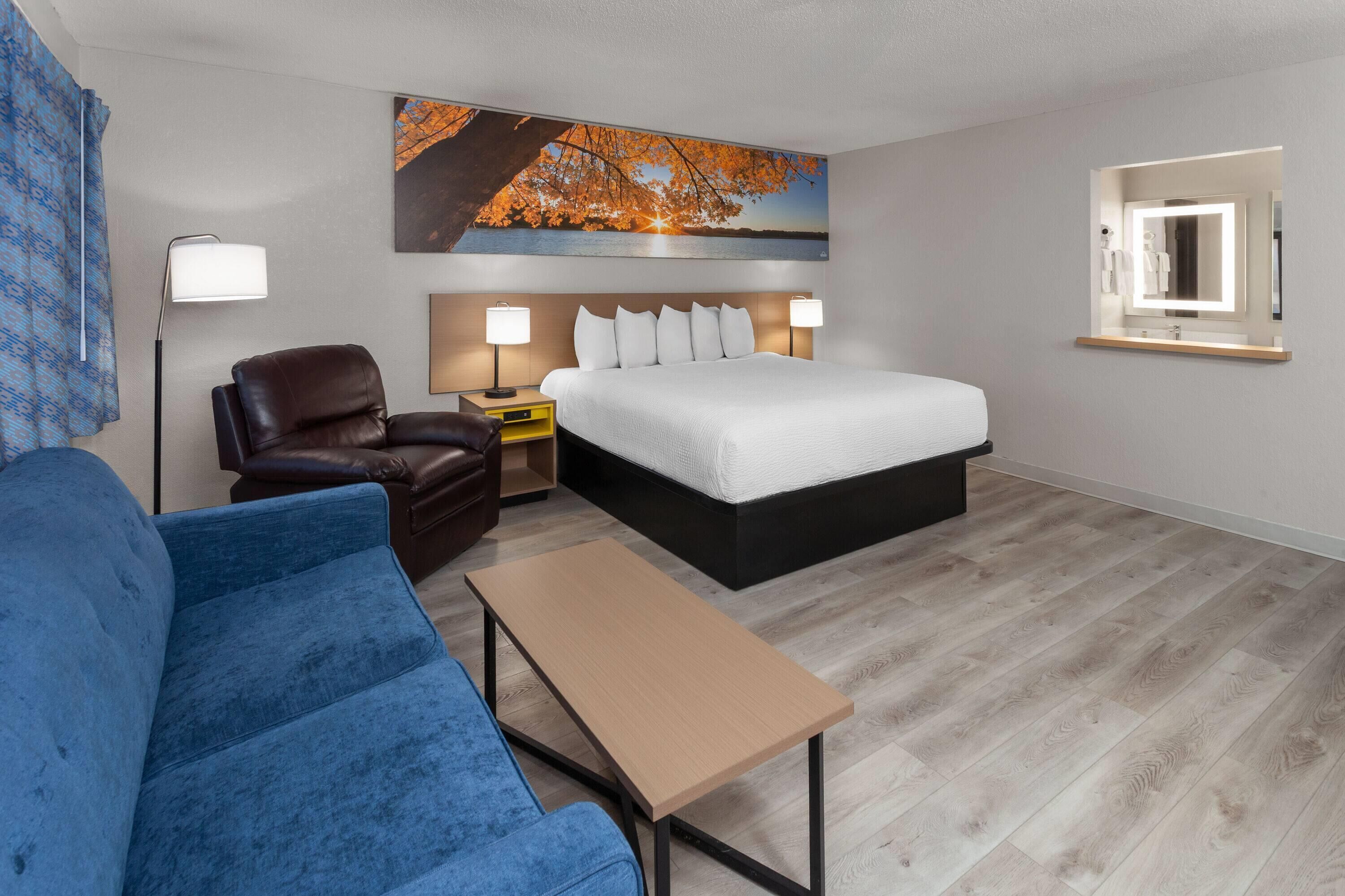 A hotel room with a bed, a blue sofa, a leather chair, a small table, a nightstand, a lamp, a painting of a sunset over water, and a mirror with a light.