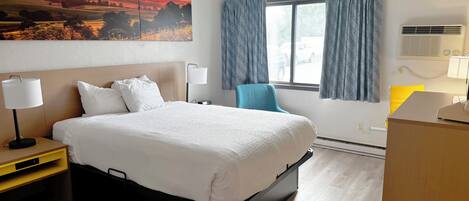A hotel room with a bed, a desk, a chair, a window with curtains, and a painting of a sunset.